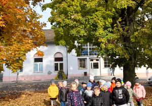 Grupa dzieci w alei kolorowych jesiennych drzew, w tle dworzec kolejowy.