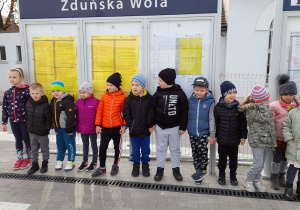 Grupa dzieci stoi na peronie, w tle rozkład jazdy pociągów.
