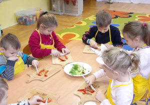Dzieci siedzą przy stoliczku i kroją warzywa.