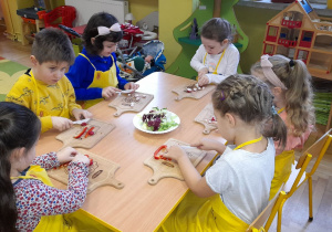 Dzieci siedzą przy stoliczku i kroją warzywa.