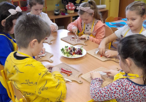 Dzieci siedzą przy stoliczku i kroją warzywa.