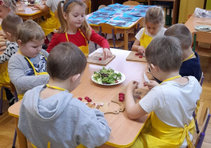 Dzieci siedzą przy stoliczku i kroją warzywa.