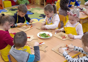 Dzieci siedzą przy stoliczku i kroją warzywa.