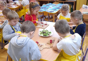 Dzieci siedzą przy stoliczku i kroją warzywa.