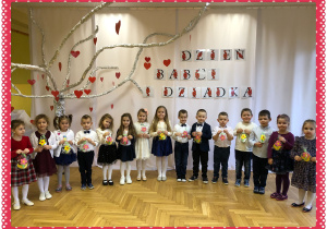 Dzieci elegancko ubrane na tle białego napisu i drzewka z sercami i napisem Dzień Babci i Dziadka.