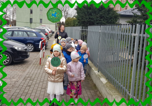 Na zdjęciu grupa dzieci przed przedszkolem. . Każde dziecko trzyma plakietkę na patyczku.