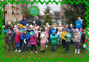 Na zdjęciu dzieci i panie w ogrodzie przedszkolnym. Dzieci trzymają plakietki dotyczące dbania o naszą planetę.
