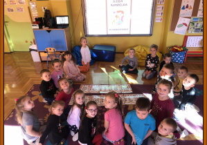 Dzieci siedzą na dywanie a na podłodze ułożony przez nie napis Dzień Kundelka, a także taki napis w tle na tablicy