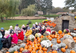 Dzieci siedzą na kamieniach, przed nimi pomarańczowe dynie i kwiaty.