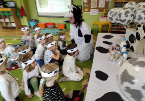 Dzieci siedzą na dywanie z opaskami w łaty krowy, słuchają opowiadania pani przebranej za krowę