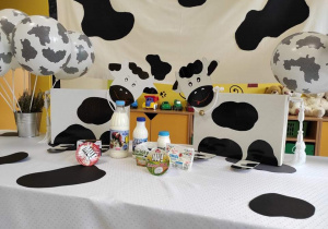 Stół ozdobiony w czarne łaty, białe balony w czarne łaty, dwie kartonowe krowy i produkty mleczne.