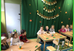 Dzieci z Lisków, Sówek, Biedronek i Jeżyków siedzą przy stolikach i biorą udział w zabawach logopedycznych. W tle zielony materiał z napisem Jesienny Konkurs Logopedyczny.