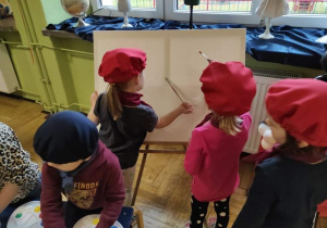 Dziewczynki w czerwonych beretach malują obraz na płótnie.