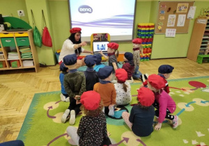 Dzieci siedzą na dywanie, chłopcy w niebieskich beretach i apaszkach, dziewczynki w czerwonych, słuchają prezentacji na temat Francji.