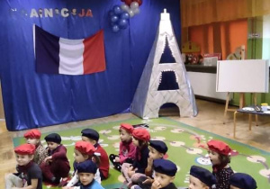 Dzieci siedzą na dywanie, chłopcy w niebieskich beretach i apaszkach, dziewczynki w czerwonych, w tle flaga Francji i wieża Eiffla.