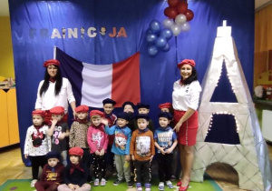 Panie i dzieci stoją na granatowym tle ozdobionym flagą Francji i wieżą Eiffla.