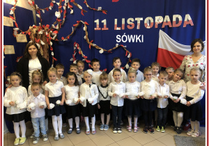Grupa dzieci wraz z Paniami w galowym stroju, w tle dekoracja patriotyczna.