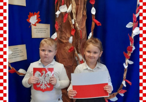 Kacper i Nadia w odświętnym ubraniu trzymają w rączkach godło i flagę Polski, w tle dekoracja okolicznościowa