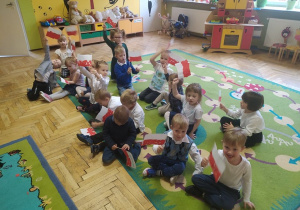 Dzieci siedzą na dywanie i machają samodzielnie wykonanymi flagami Polski.