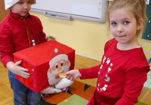 Chłopiec trzyma skrzynkę z postacią Mikołaja. Dziewczynka karmi Mikołaja ciasteczkami.