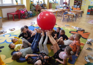 Dzieci leżą na dywanie, stopami podnoszą do góry piłkę z kolcami.