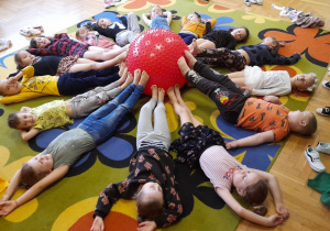 Dzieci leżą na dywanie, stopy opierają o piłkę z kolcami.