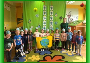 Dzieci z panią Olą z wykonaną przez siebie pracą plastyczną na tle zielonego materiału z napisem Dzień Ziemi.