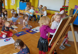Dzieci malują farbami. Na pierwszym planie dziewczynki stoją przy sztaludze, malują.