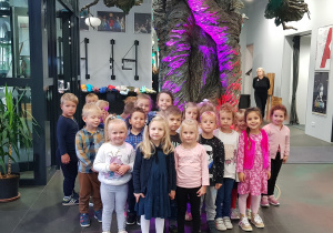 Na zdjęciu grupa dzieci w teatrze. Za nimi duże, kolorowe drzewo.