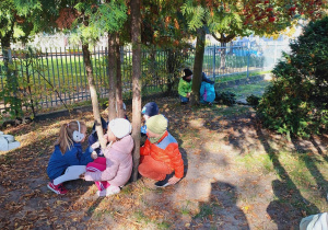 Dzieci kucają pod drzewem w ogrodzie przedszkolnym.