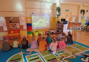 Dzieci siedzą na dywanie, oglądają film edukacyjny.
