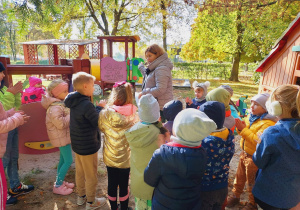 Dzieci wraz z panią obserwują drzewa w ogrodzie przedszkolnym.