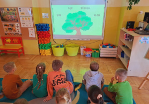 Dzieci siedzą na dywanie i oglądają na tablicy multimedialnej film edukacyjny dot.drzew.
