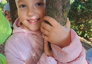 Dziewczynka przytula się do drzewa w ogrodzie przedszkolnym.