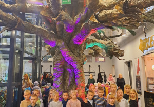 Dzieci pozują do zdjęcia przed magicznym kolorowym drzewem w budynku teatru.