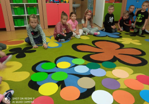 Dzieci siedzą na dywanie, a po środku kolorowe kropki wycięte z papieru.