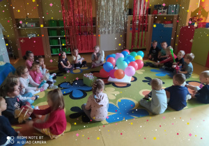 Dzieci siedzą w kole na dywanie, po środku balony i upominki.