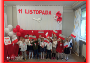 Dzieci na tle biało - czerwonej dekoracji 11 Listopada, w rączkach trzymają flagi Polski.