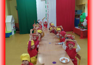 Dzieci siedzą przy stolikach, w strojach kucharskich, w ręku trzymają wałek do ciasta, w tle flaga Włoch.
