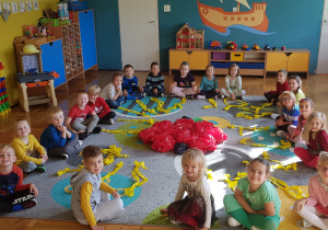 Grupa dzieci siedzi na dywanie. Przed każdym dzieckiem leżą paski żółtej bibuły. W środku , na dywanie, leży duże jabłko z czerwonej folii.