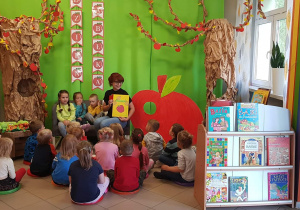 Na zdjęciu grupa dzieci. W tle napis Dzień Jabłka. Pani czyta bajkę o jabłuszku.