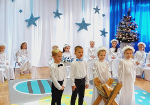 Dzieci w odświętnych strojach stoją na tle świątecznej dekoracji. Dwie dziewczynki i dwóch chłopców śpiewają piosenkę.