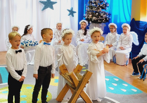 Dzieci w odświętnych strojach stoją na tle świątecznej dekoracji. Dwie dziewczynki i dwóch chłopców śpiewają piosenkę.