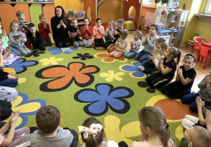 Dzieci siedzą na dywanie i naśladują pokazywane przez panią gesty