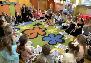 Dzieci siedzą na dywanie i naśladują pokazywane przez panią litery