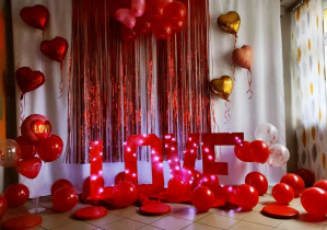 Walentynkowa dekoracja holu przedszkolnego. Białe tło, czerwona kurtyna, czerwone balony i duży czerwony napis love.