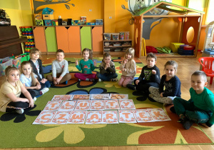 Dzieci z grupy 5 siedzą na dywanie wokoło ułożonego napisu "tłusty czwarte"k
