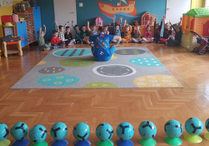 Na pierwszym planie niebieskie piłki. Dzieci siedzą na podłodze, podnoszą ręce.