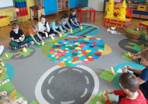 Dzieci siedzą na dywanie, robią wielkanocne kurki z papieru.