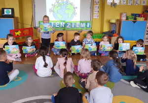 Dzieci trzymają obrazki. W tle napis Dzień Ziemi i obrazek przedstawiający Ziemię.